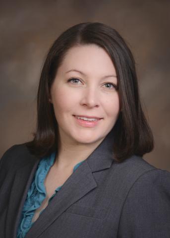 Headshot of Nicole. She has medium length brown hair. She is wearing a grey suit jacket and a blue top. She is smiling. 