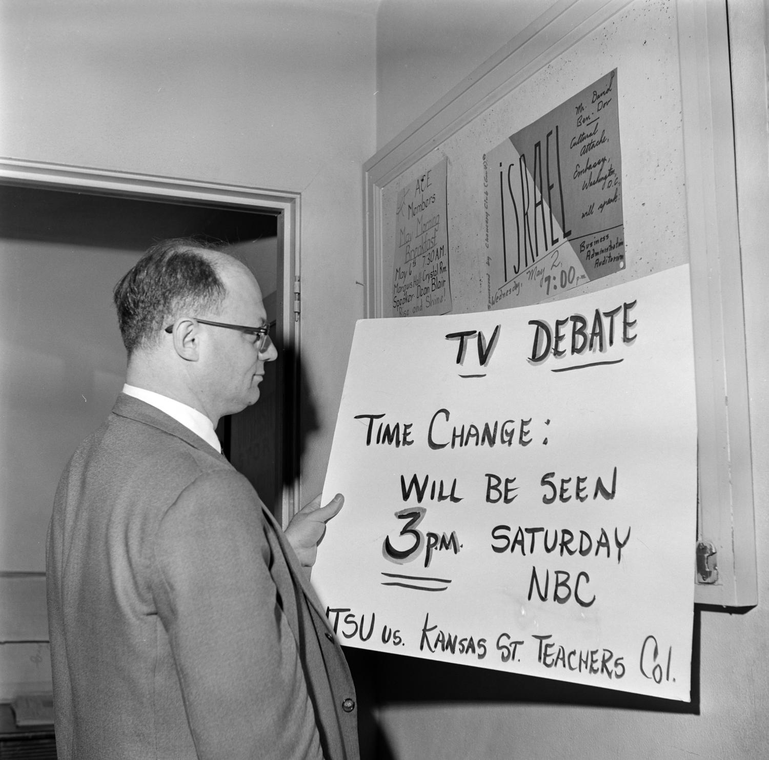 Dr. DeMougeot in front of a card noting a TV debate will be seen at 3 PM Saturday on NBC