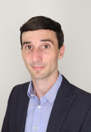 Andrew Huebner in a suit standing in front of a gray background.