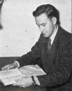 Photograph of Thomason sitting down, reading a book placed on a table, from the left profile view