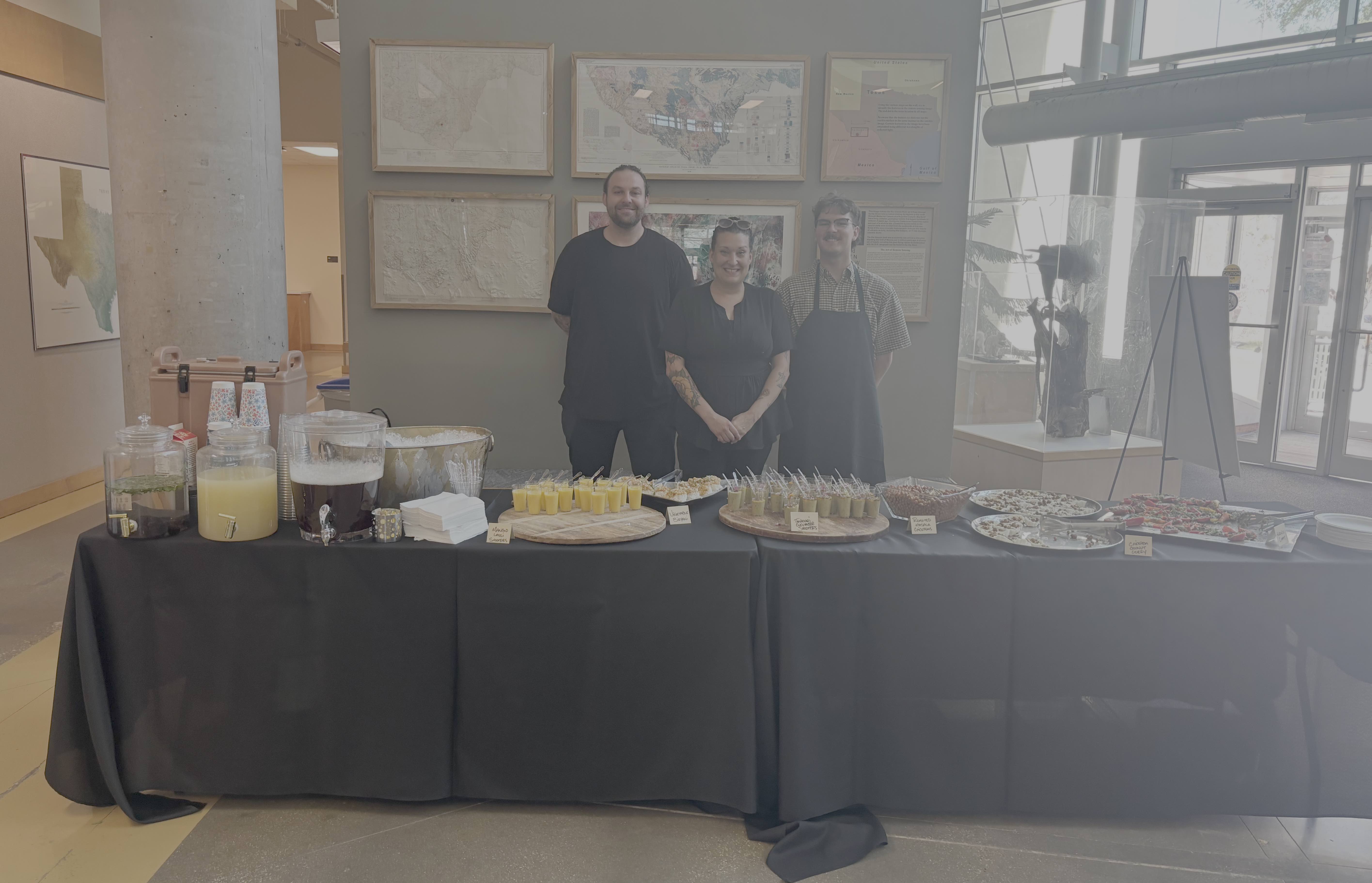 Three people stand behind a table full of food.