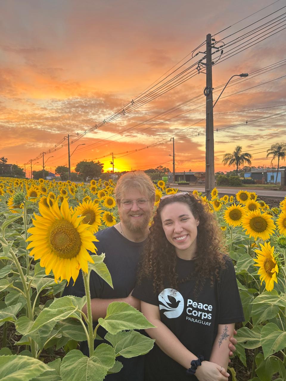 Delaney and her partner, Lee, pose for a photo in a sunflower field with a bright orange sunset behind them.