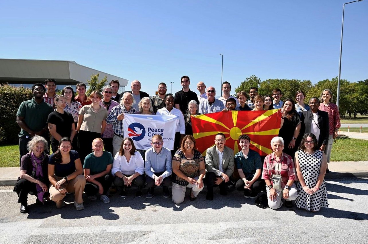 A group of Peace Corps Volunteers and Host Country Nationals pose with a Peace Corps flag and the flag of Macedonia.