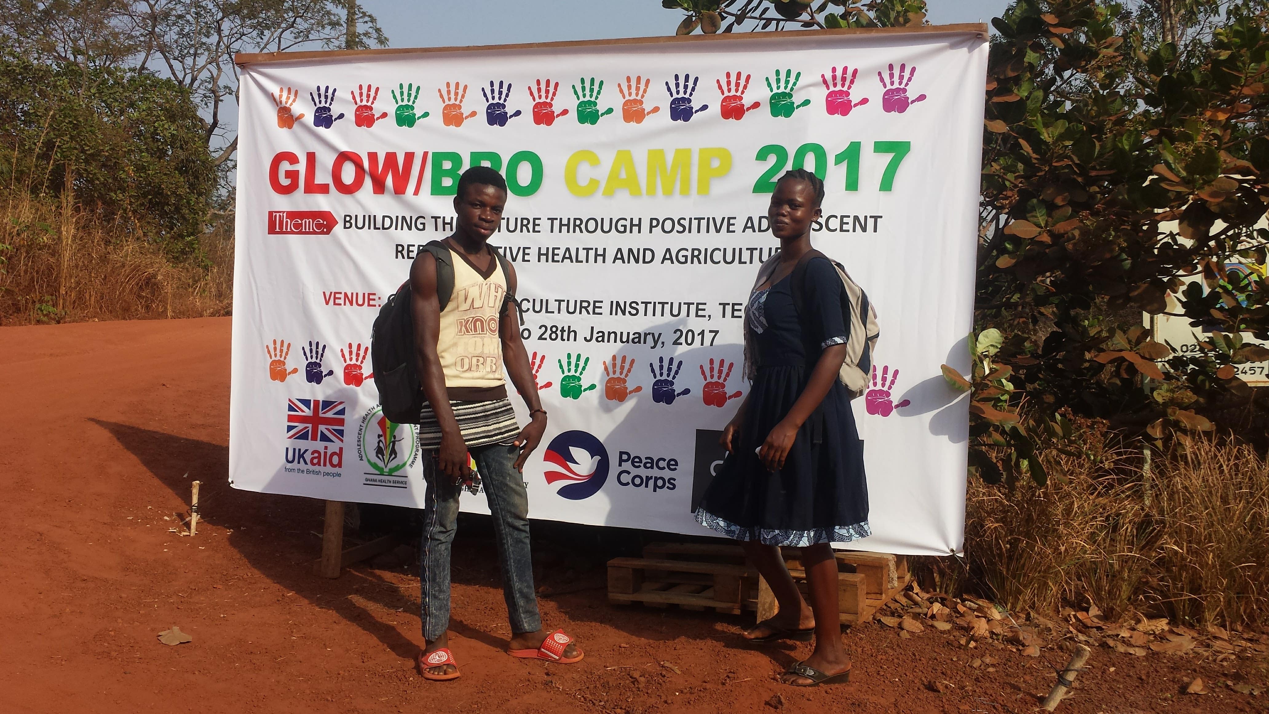 Two locals from Ghana stand in front of a large poster for a 2017 youth camp.