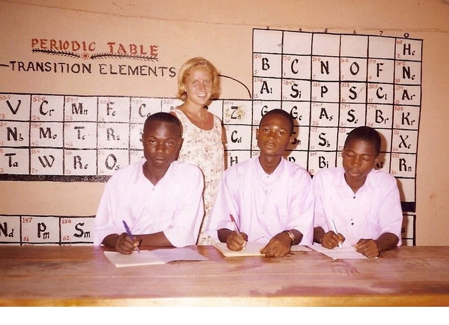 Lura stands in front of a Periodic Table of Elements that she painted. Three students sit in front of her.
